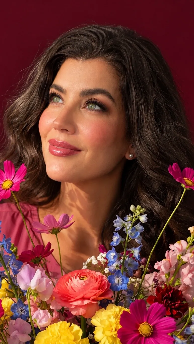 Christy gazing upward behind a vivid bouquet of cosmos, ranunculus, delphinium and marigolds against a burgundy backdrop.