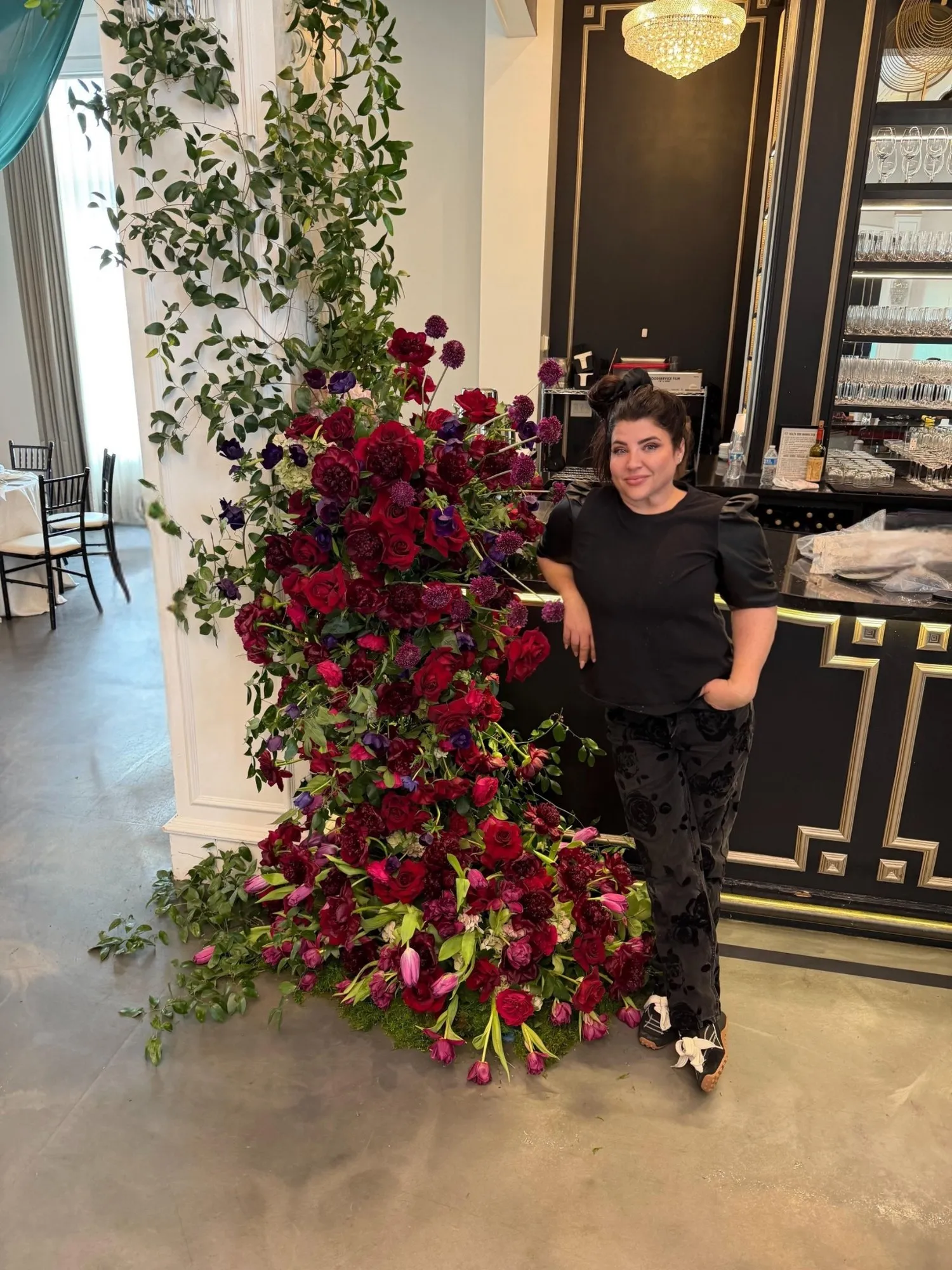 Christy beside a floor-to-ceiling installation of red roses and trailing greenery.