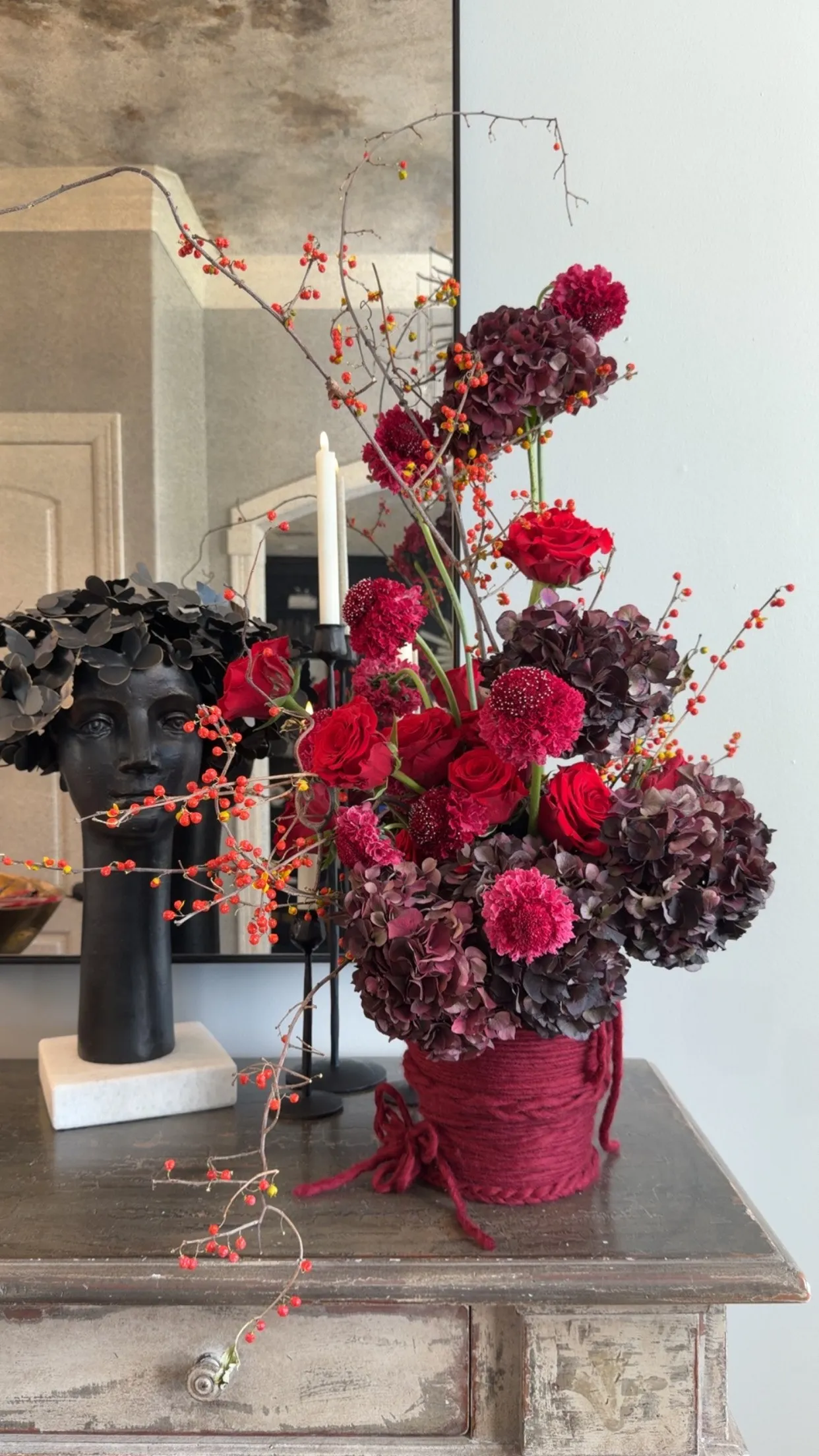 Burgundy hydrangea, red roses and bittersweet branches in a yarn-wrapped vessel on an antique console beside a black bust.
