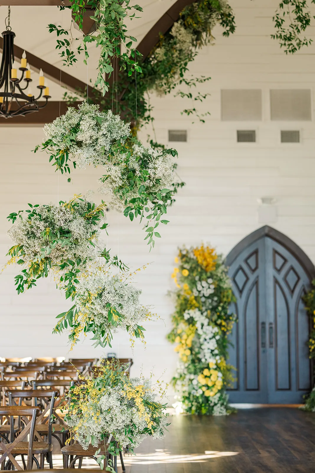 Clouds of baby's breath and greenery suspended over a chapel aisle, with a yellow and white floral column beside arched blue doors.