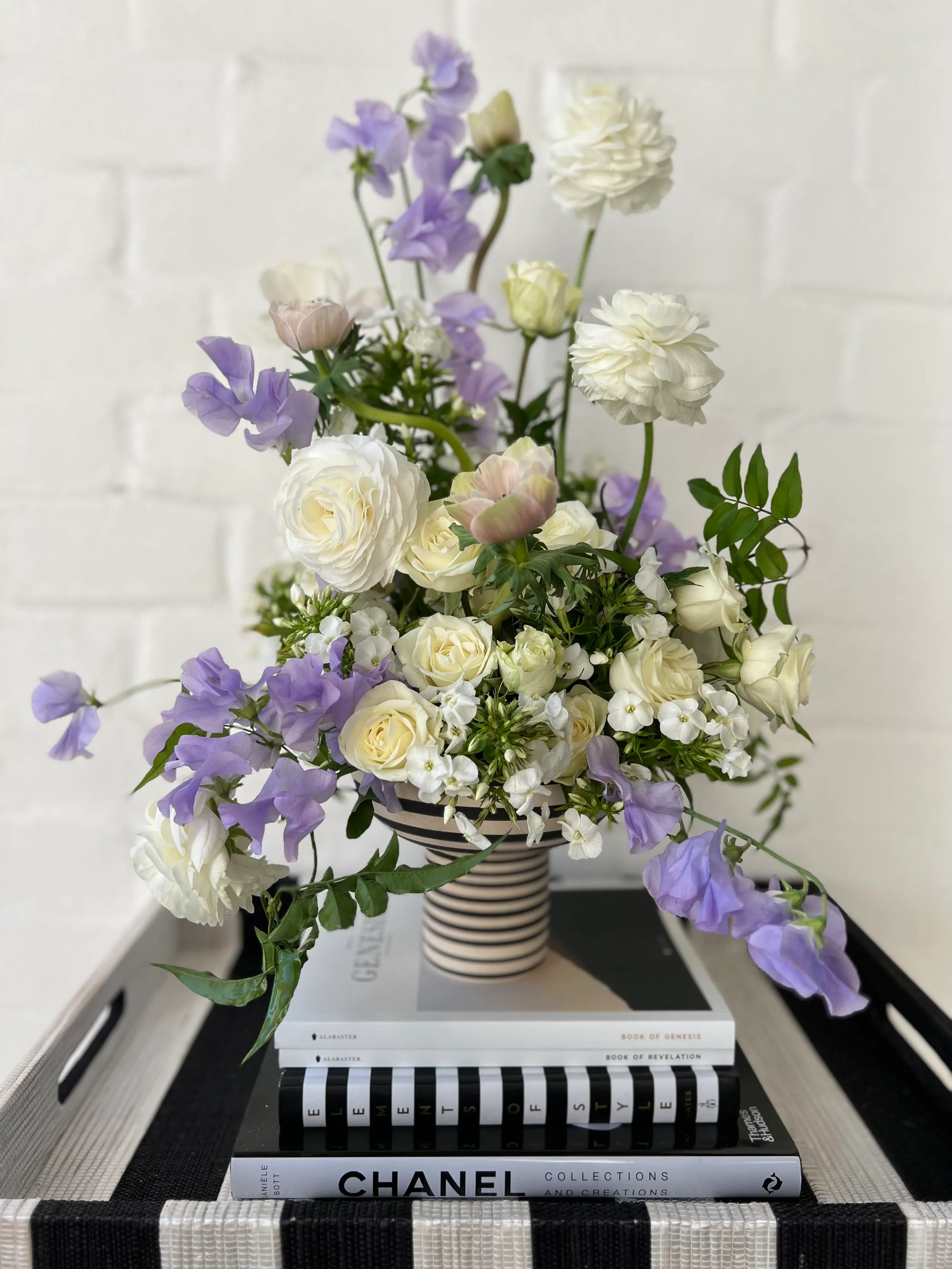 Layered arrangement of white ranunculus, lilac sweet peas and greenery in a striped vase, styled on a stack of design books.