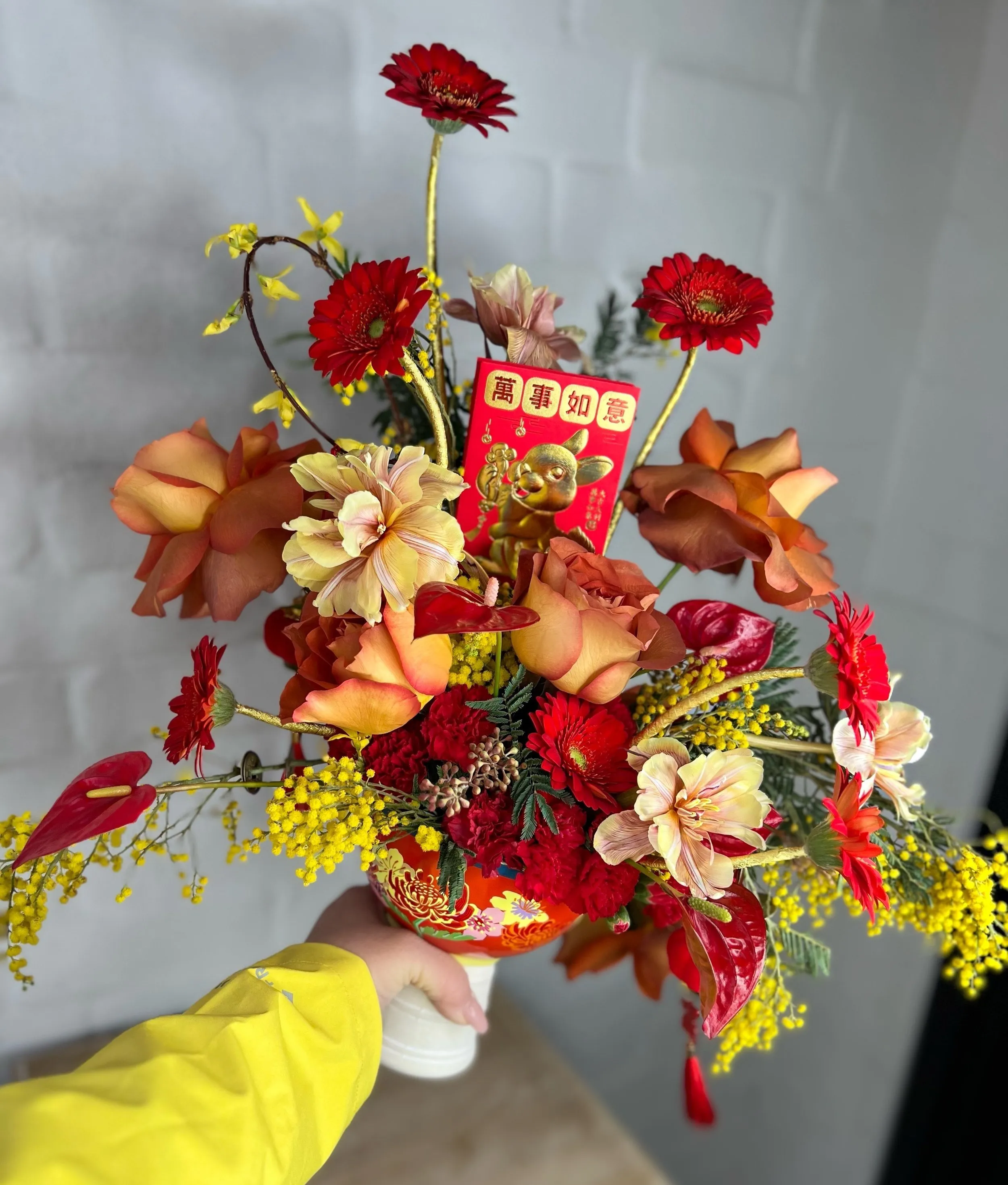 Hand-held celebration bouquet of red dahlias, orange calla lilies and yellow accents against a white brick wall.