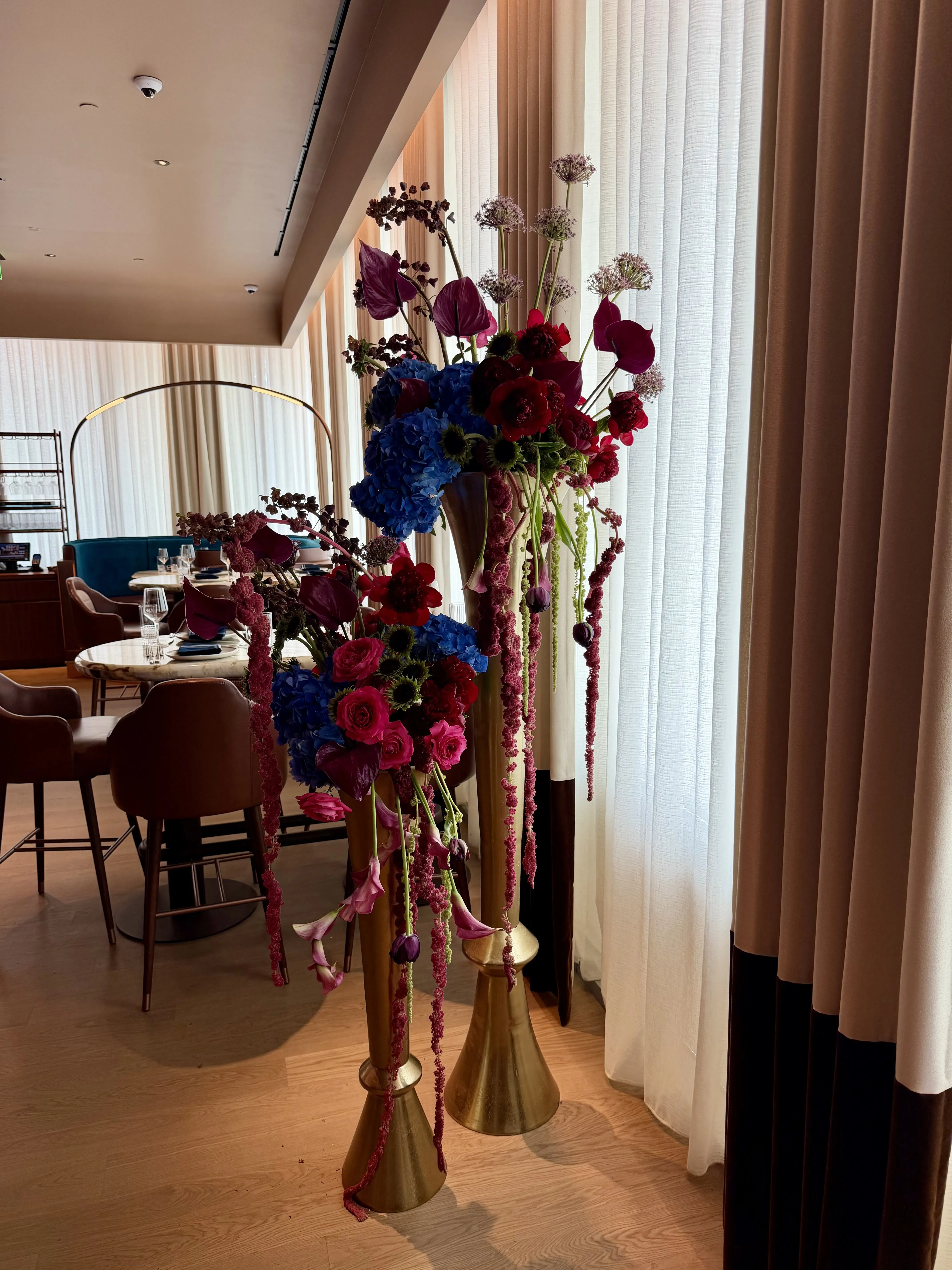 Twin brass pedestal vessels holding towering red, fuchsia and blue arrangements with trailing amaranthus.