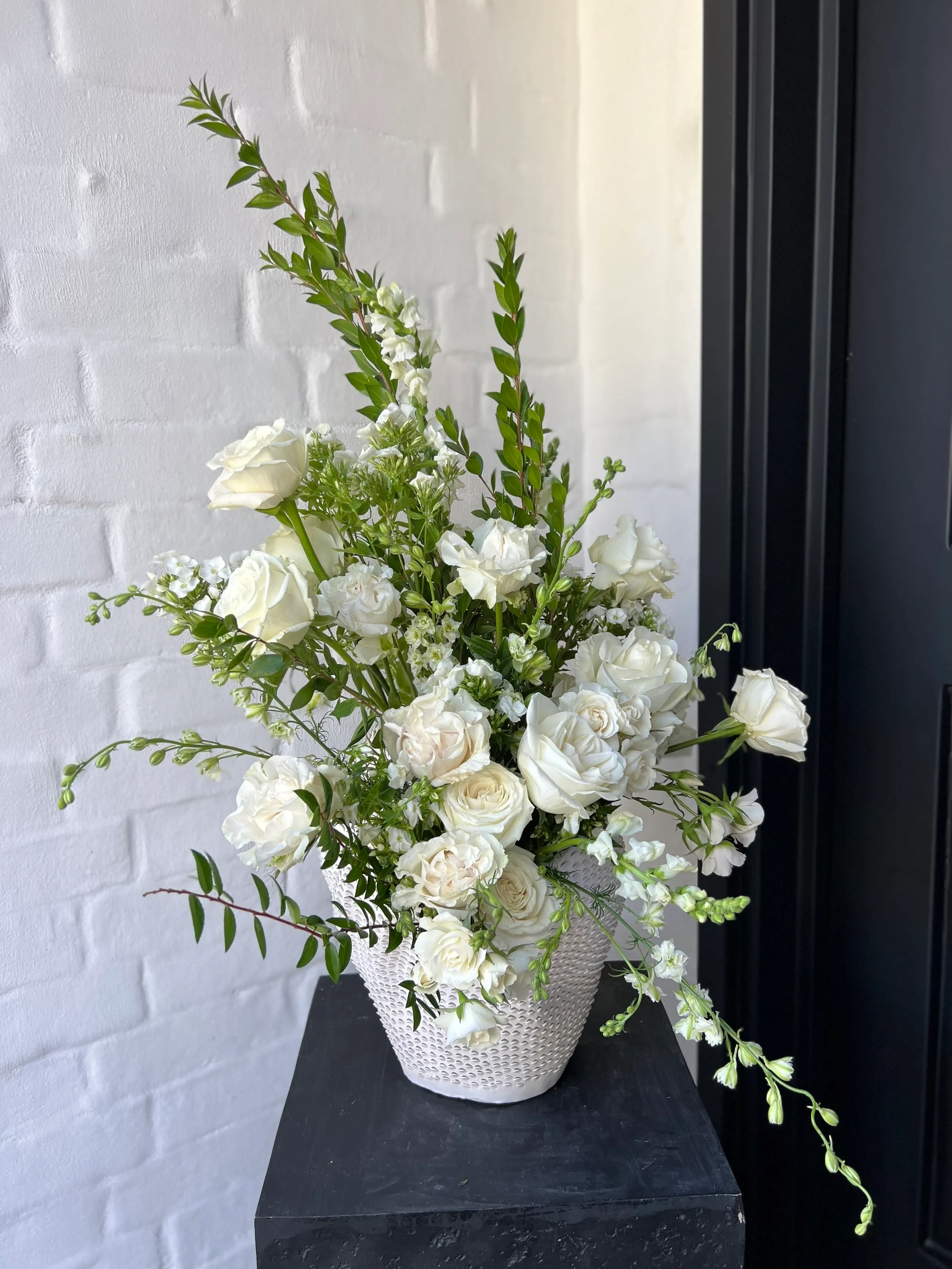 White and green floral arrangement with roses and trailing greenery in a textured white urn, set against a white brick wall.