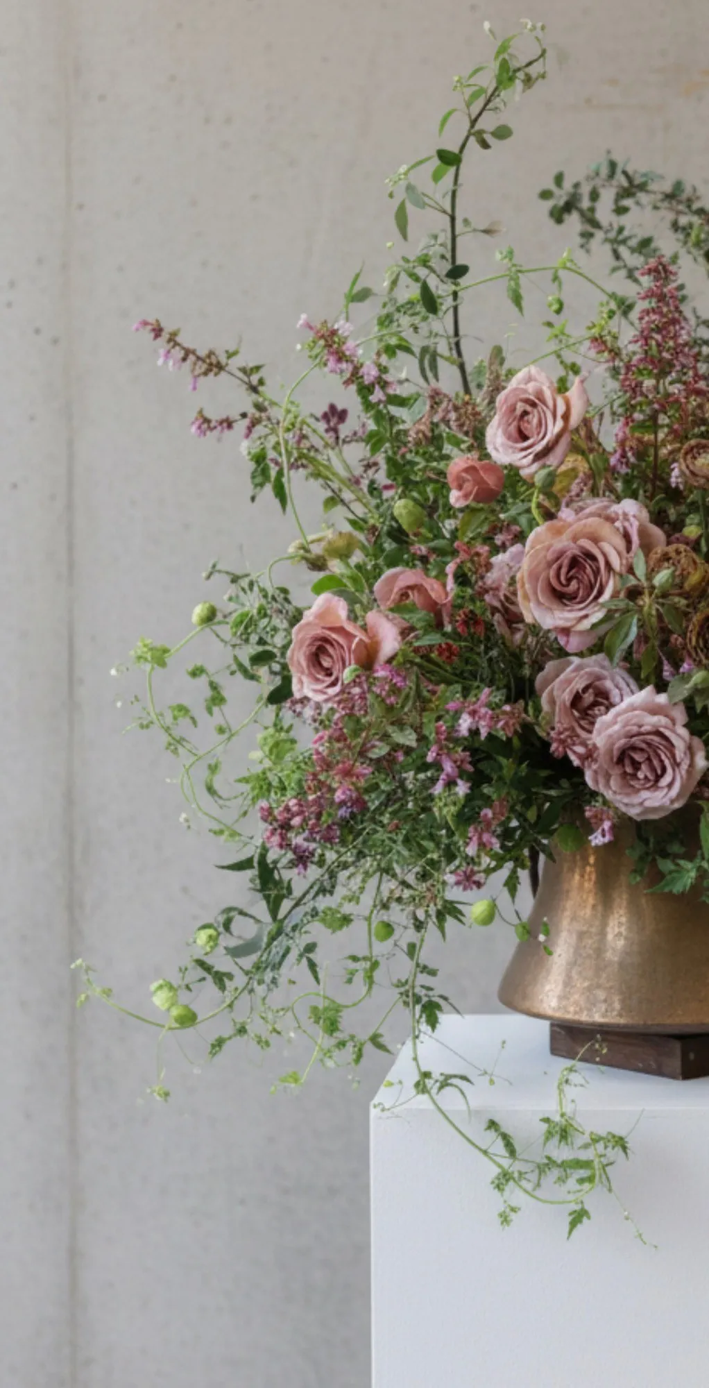 Loose garden arrangement of mauve roses and trailing greenery in a bronze vessel on a white plinth.