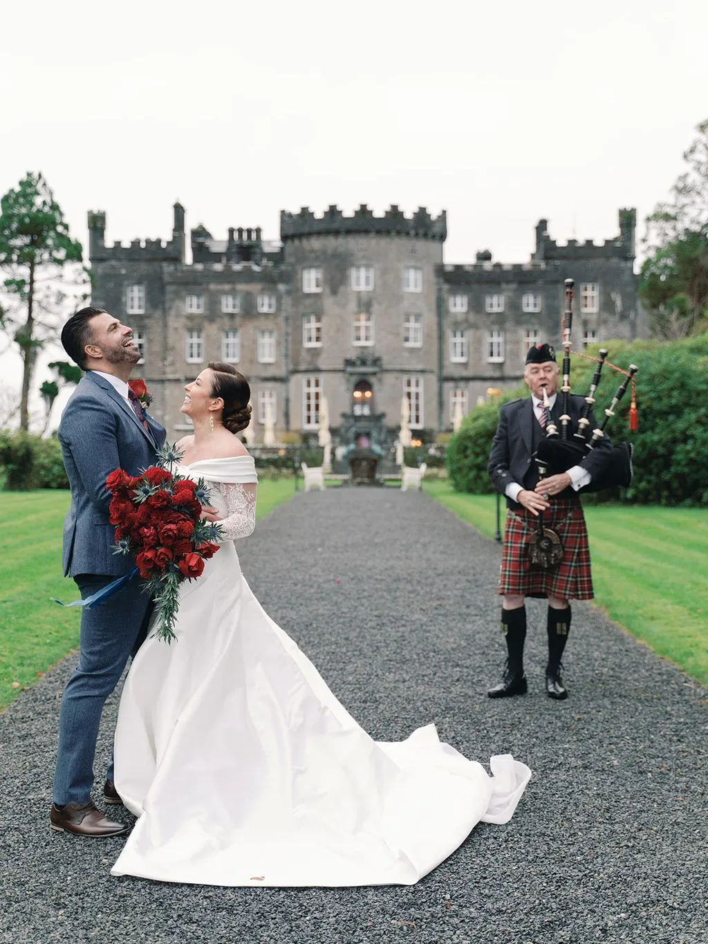 Newlyweds laughing with a red bouquet while a bagpiper plays before an Irish castle.