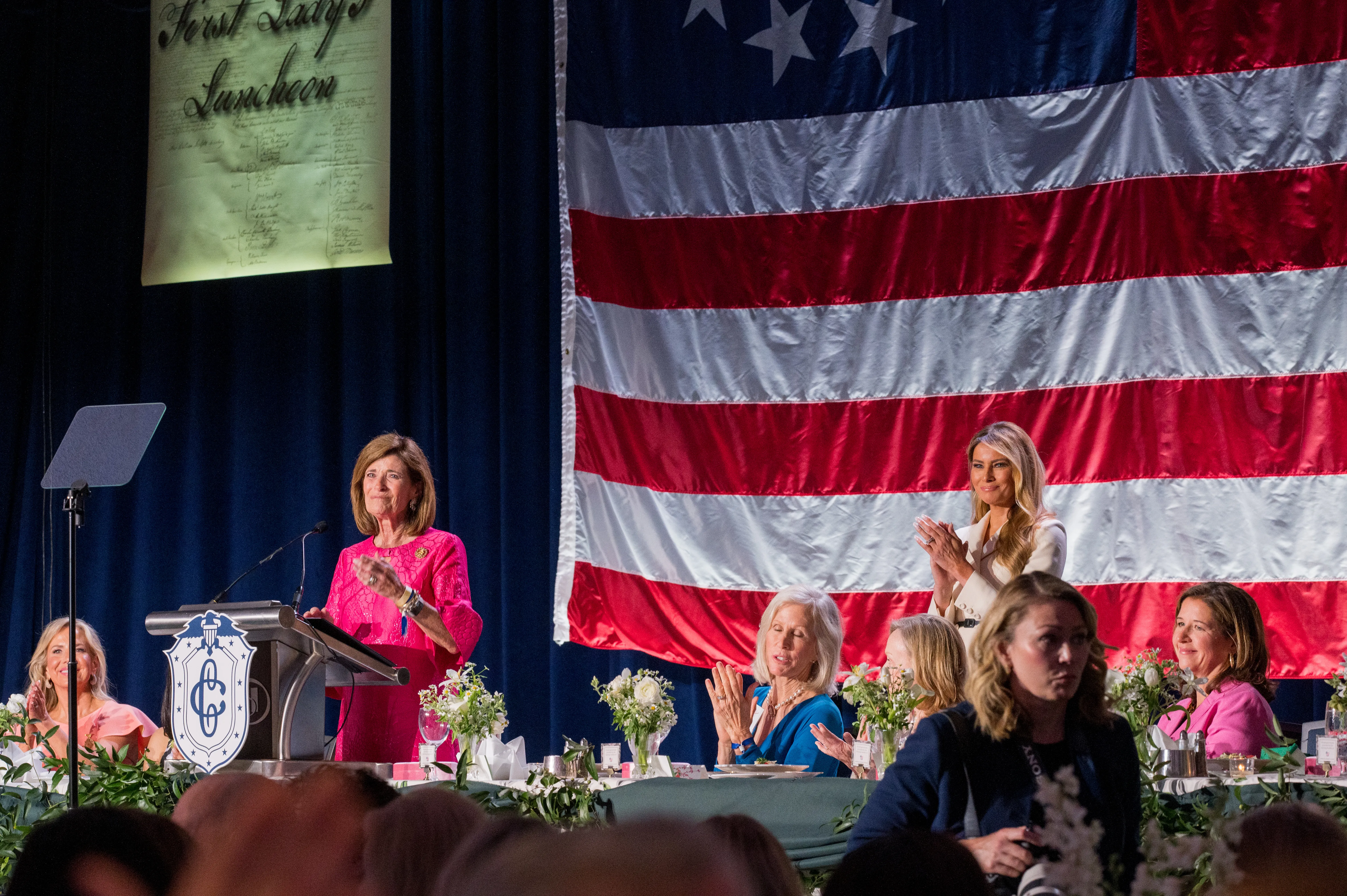 Speakers on stage before an American flag at the First Lady's Luncheon, tables dressed in white florals.