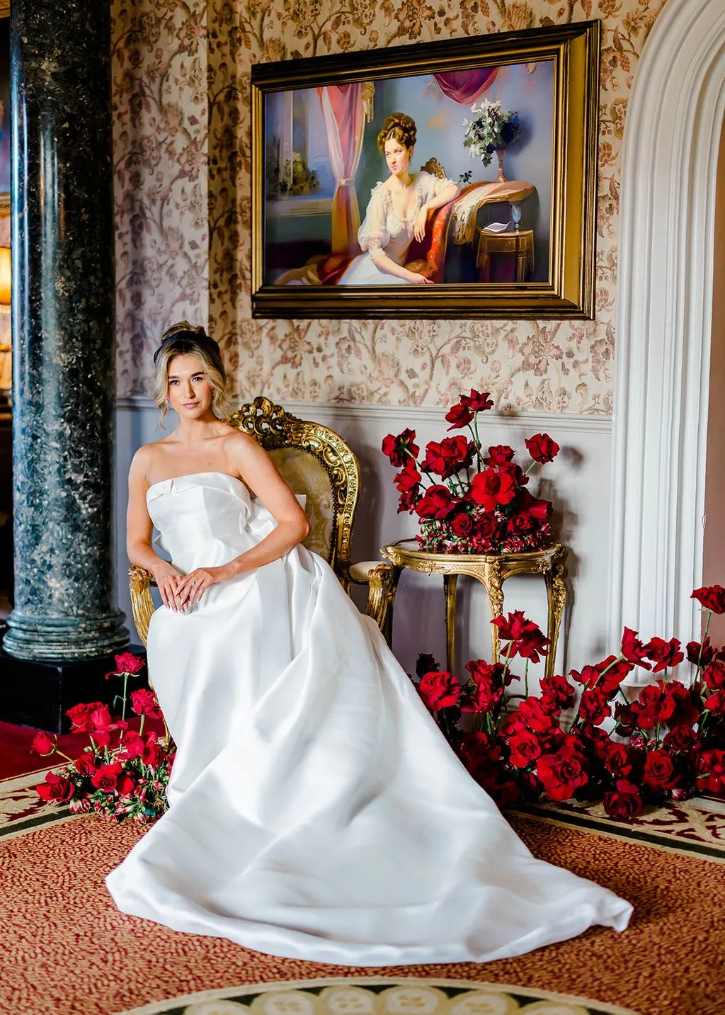 Bride in a gilt chair surrounded by red roses beneath an oil portrait.