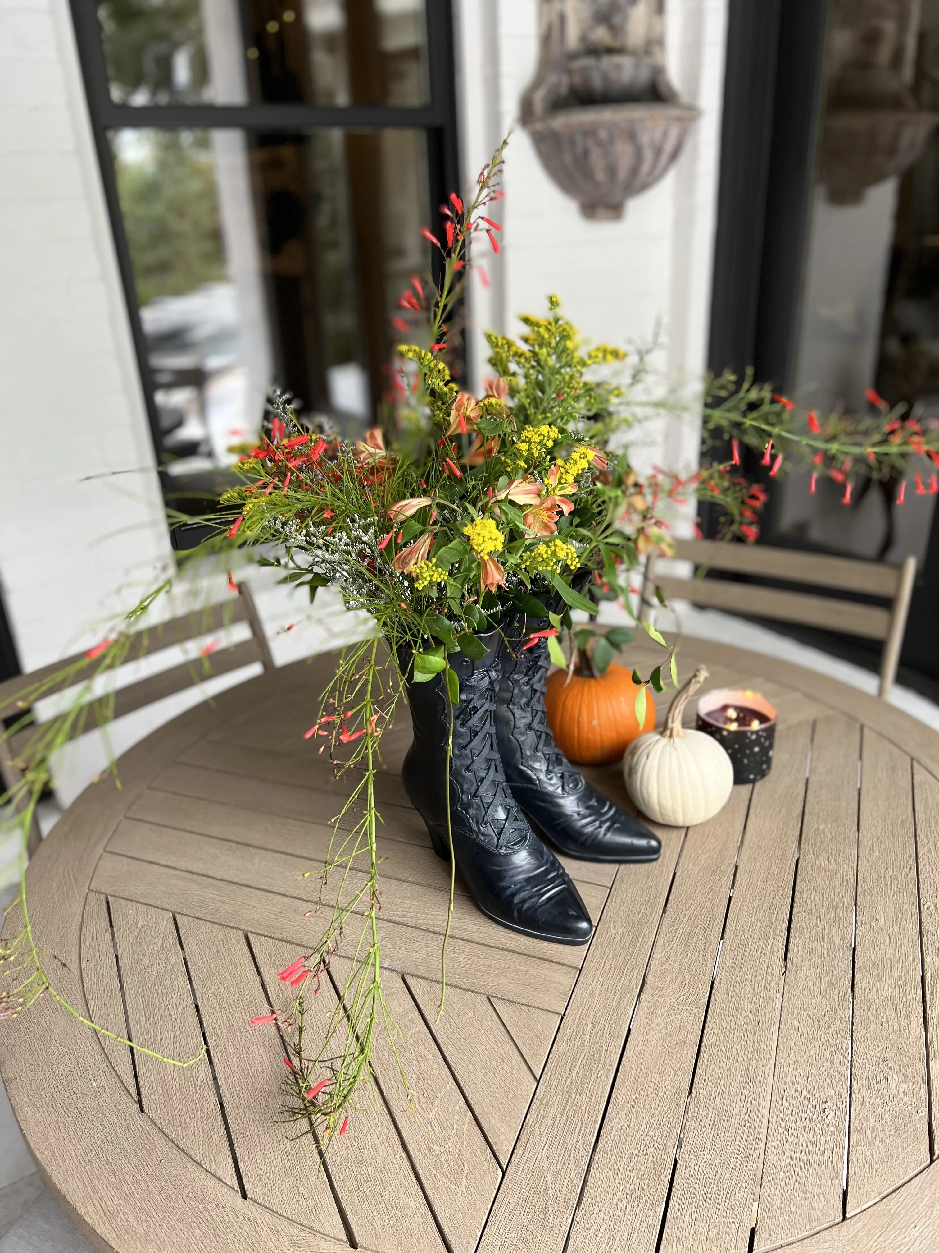 Autumn porch vignette: fall florals arranged in black cowboy boots beside pumpkins on a round wooden table.