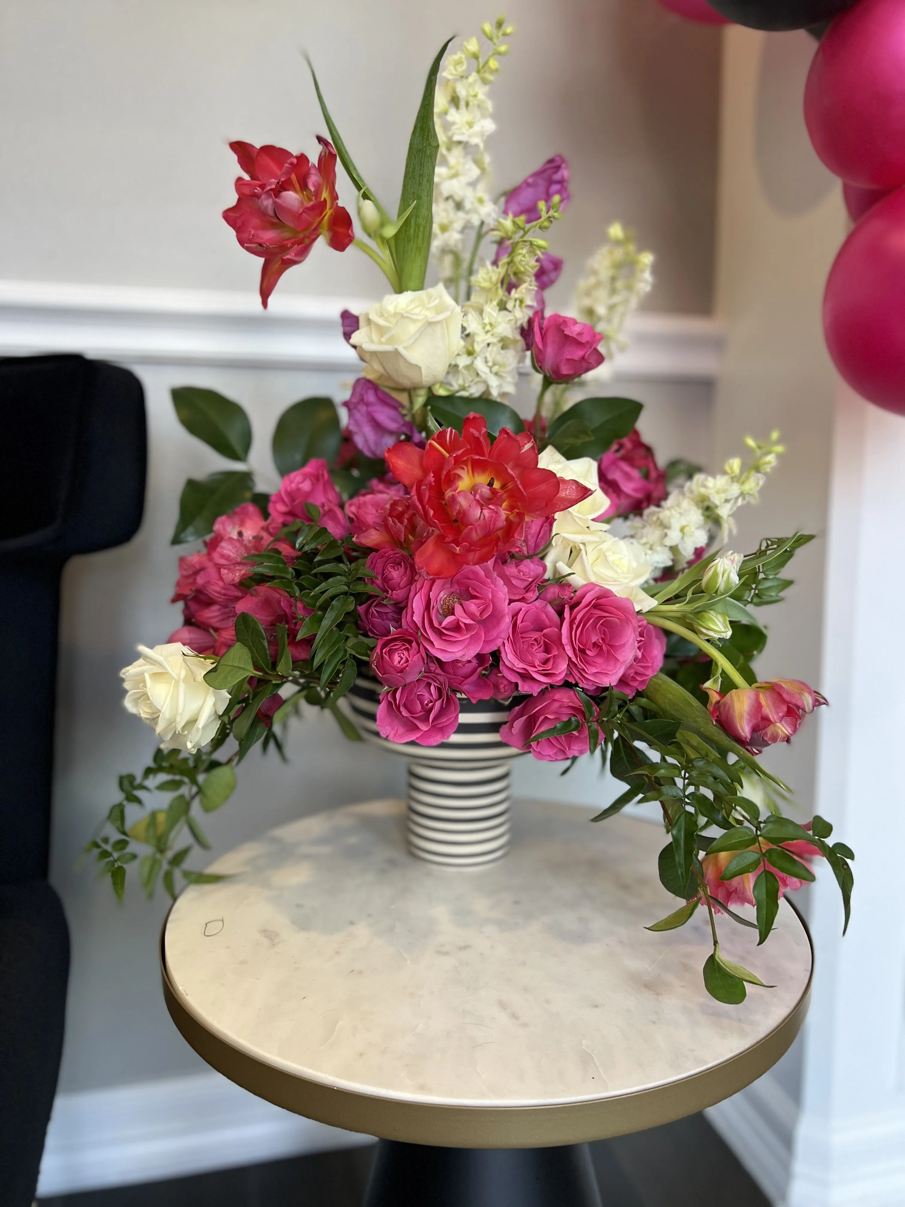Fuchsia spray roses, red parrot tulips and white stock arching from a striped compote on a marble side table.