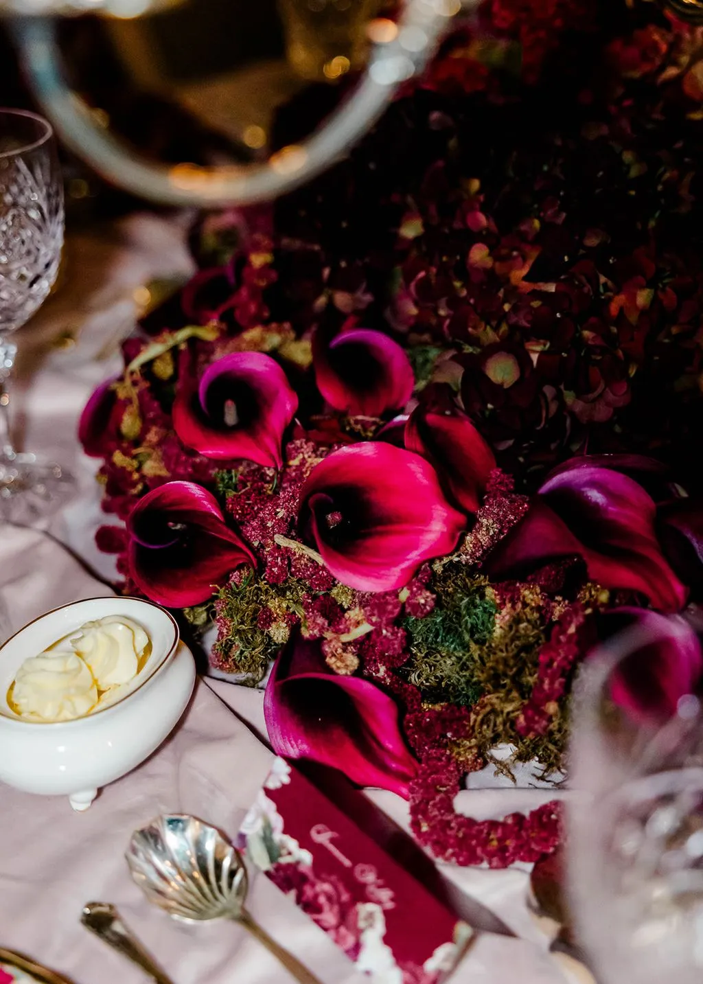 Candlelit tablescape detail with magenta calla lilies, burgundy hydrangea and moss beside crystal and silver.