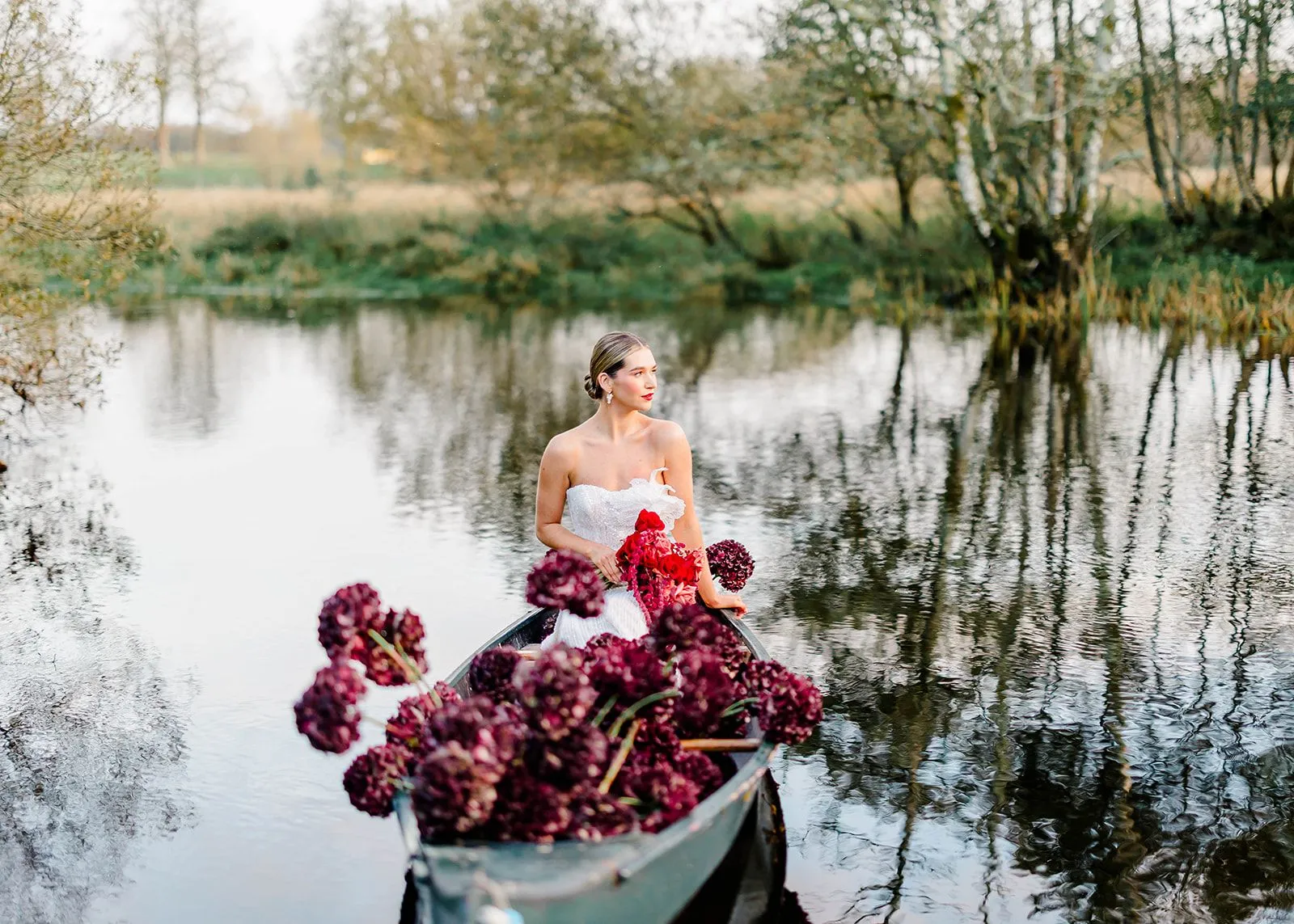 Bride seated in a canoe filled with deep burgundy hydrangeas on a still pond at dusk.