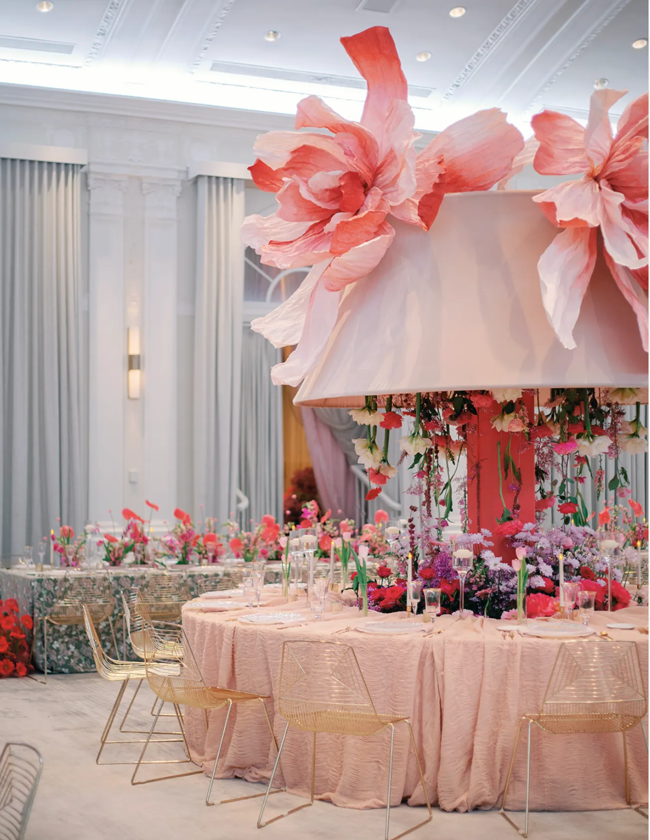 Ballroom table beneath an oversized blush lampshade crowned with giant paper flowers, hung with trailing fresh florals, ringed by gold wire chairs.