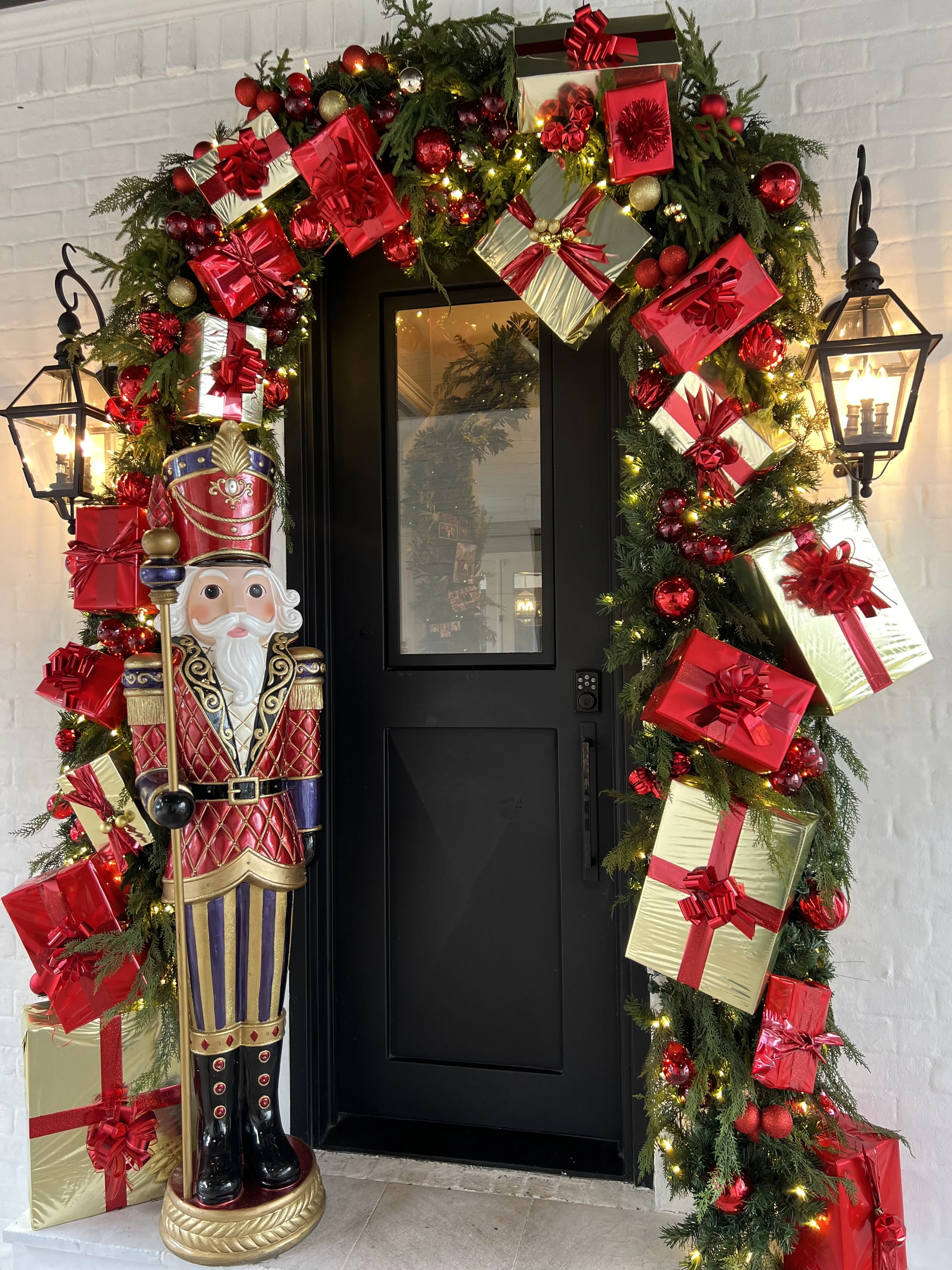 Black front door arched with garland, red bows and wrapped gifts, flanked by a life-size nutcracker.