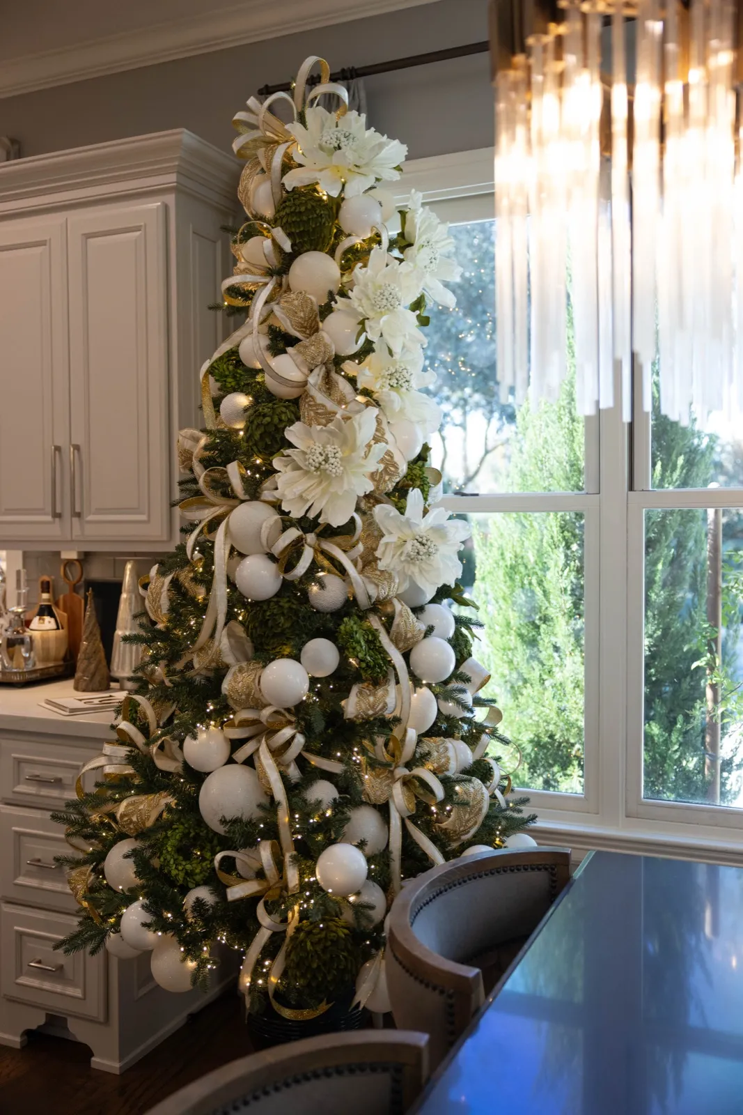 Tree dressed in white magnolias and oversized ornaments beside a bright kitchen window.