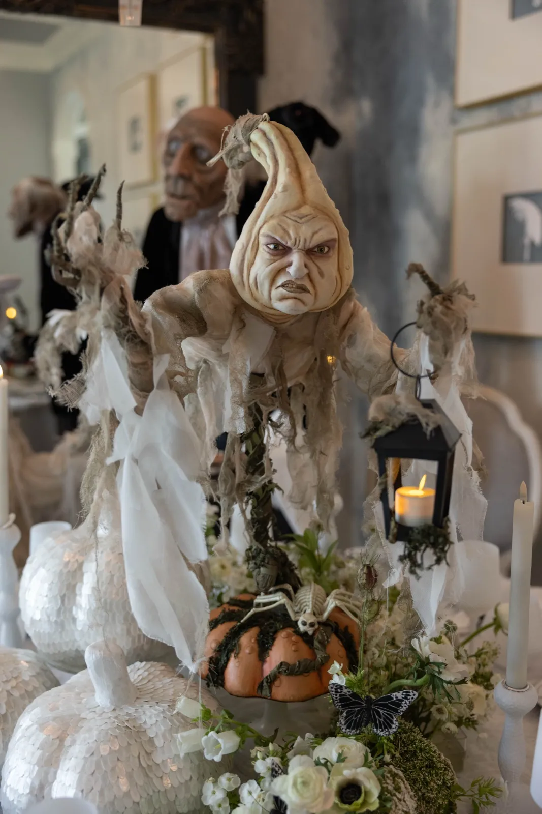 Gourd-headed jester figure with a lantern styled among white pumpkins and moss.