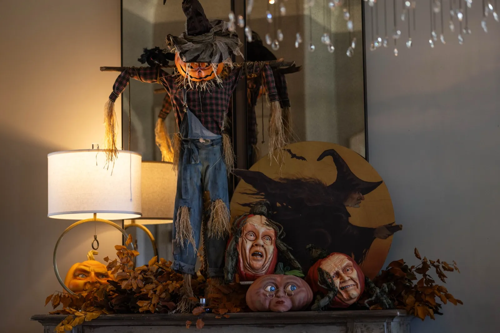 Scarecrow with a crow perched over a mantel of autumn foliage and carved-head accents.