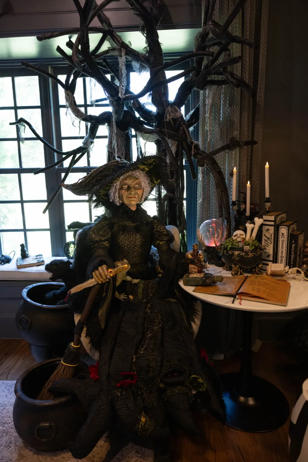 Life-size witch with cauldrons and a crystal ball beneath a black branch tree in a breakfast nook.